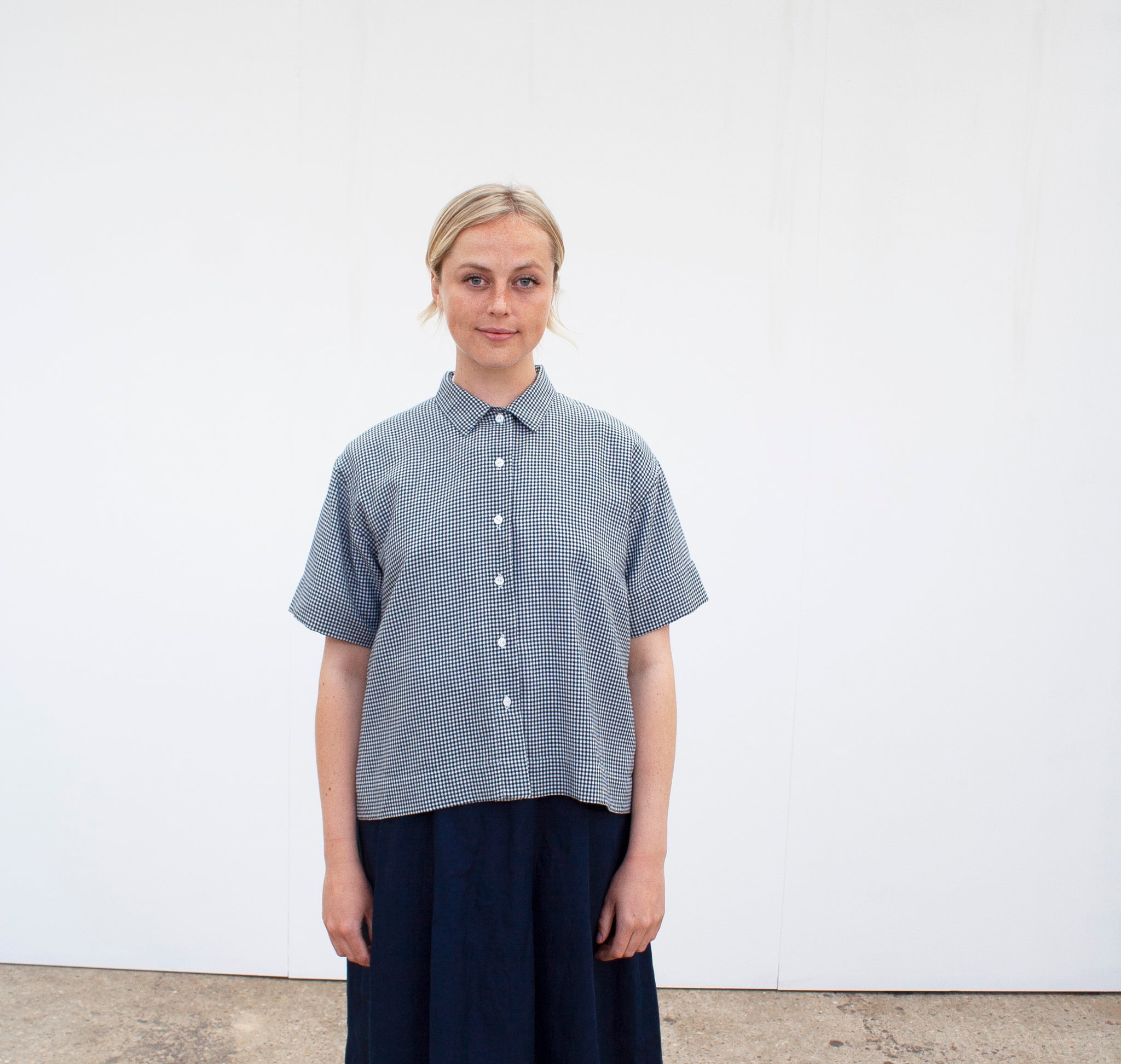 a blonde woman looking straight ahead in a white space wearing a short sleeved gingham shirt