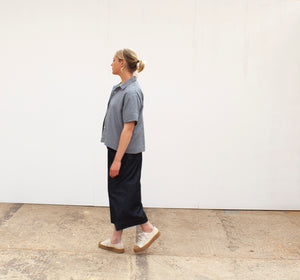 A young blond woman side view wearing a ging=ham short sleeved shirt and a navy linen skirt