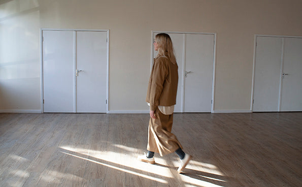 young blond woman standing in a sy=unlit room, wearing a tan cotton casual jacket and trousers