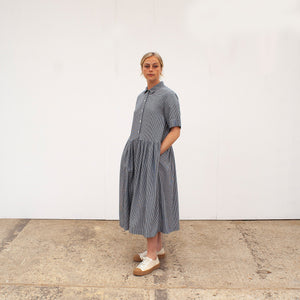 woman stands in front of a white wall, her hands in her pockerts, wearing a gingham dress