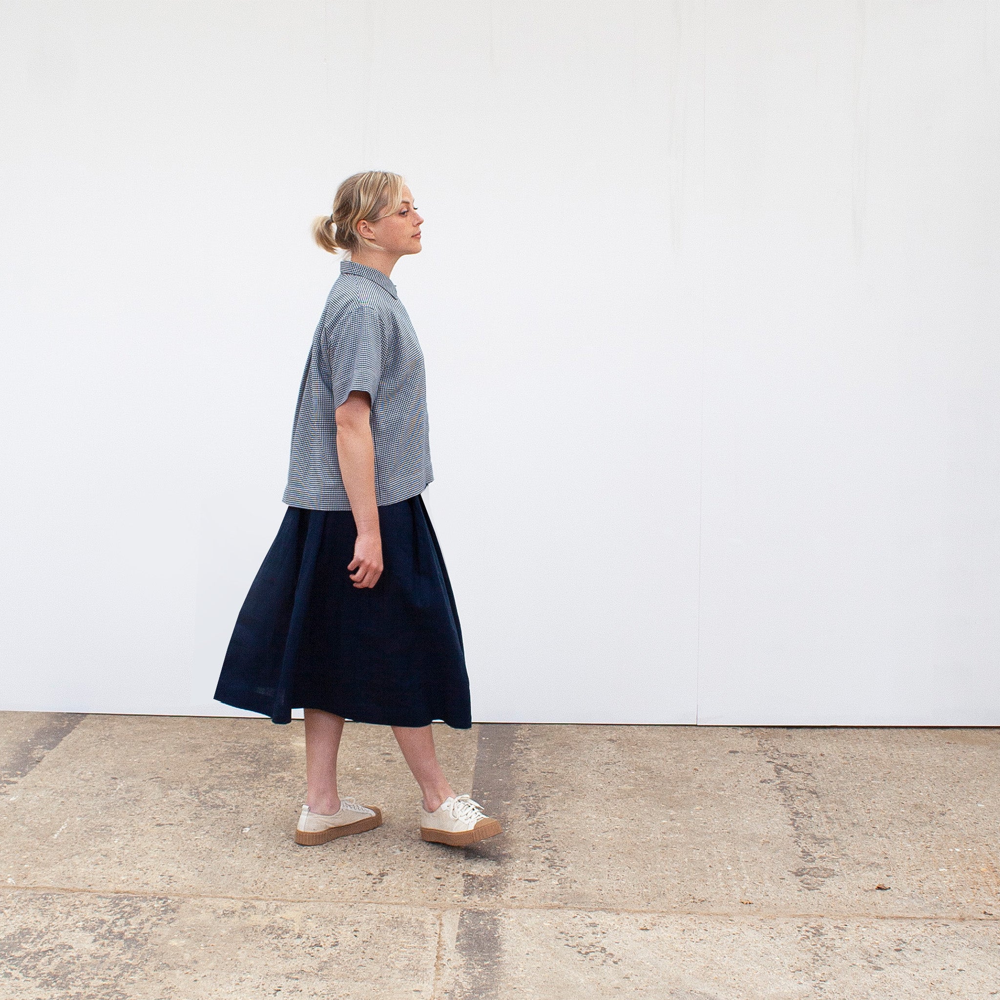 side view of A young blond woman facing the camera wearing a ging=ham short sleeved shirt and a navy linen skirt