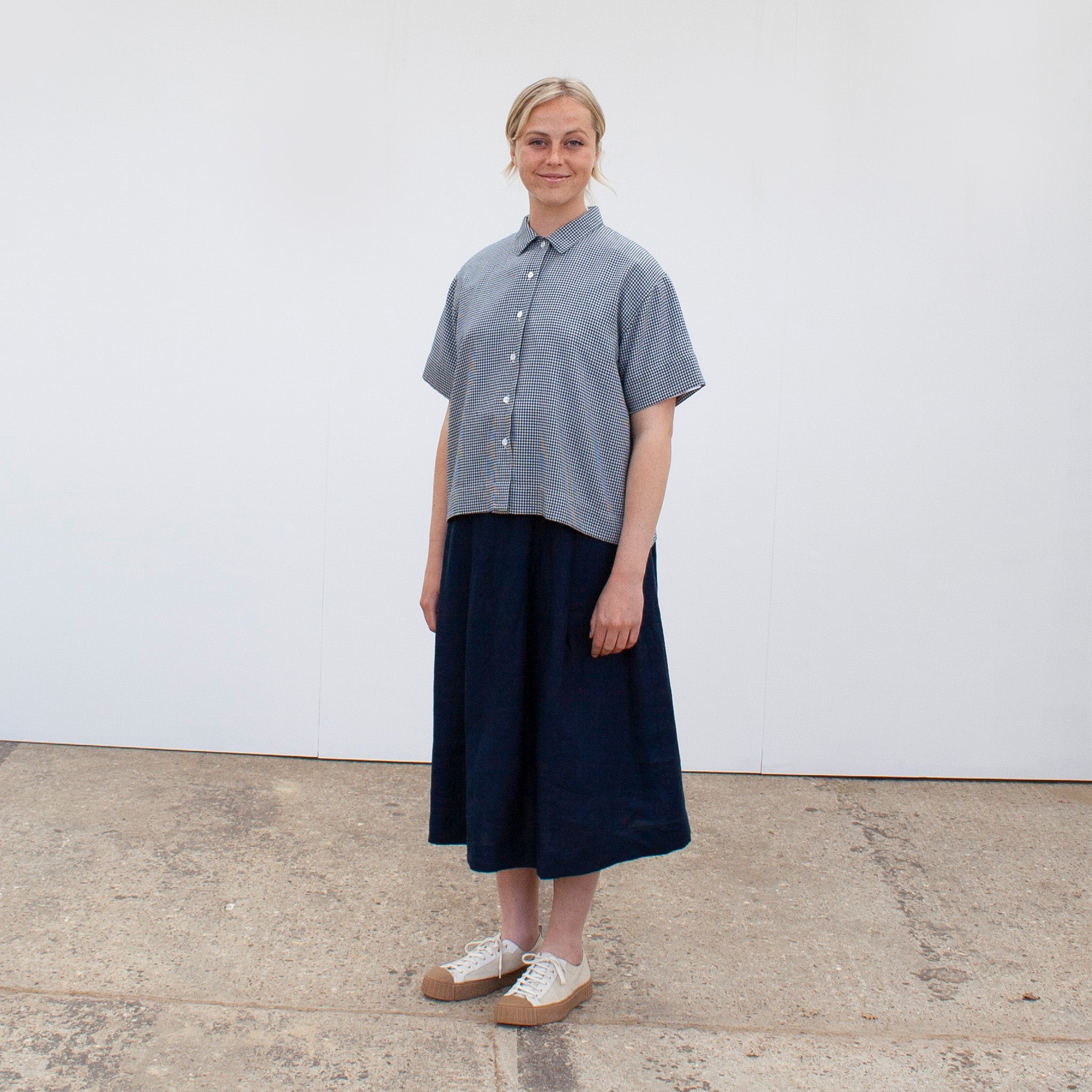 A young blond woman facing the camera wearing a ging=ham short sleeved shirt and a navy linen skirt