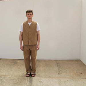 A young man stands still, facing forward, wearing a tan waistcoat and tan chore trousers made by Pajotten