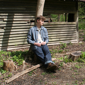 a young man sits outside on a sunny day wearing simple denim chore wear