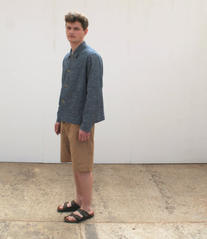 A young man stands in a white space wearing a light blue denim chore jacket and a pair of tan coloured cotton shorts