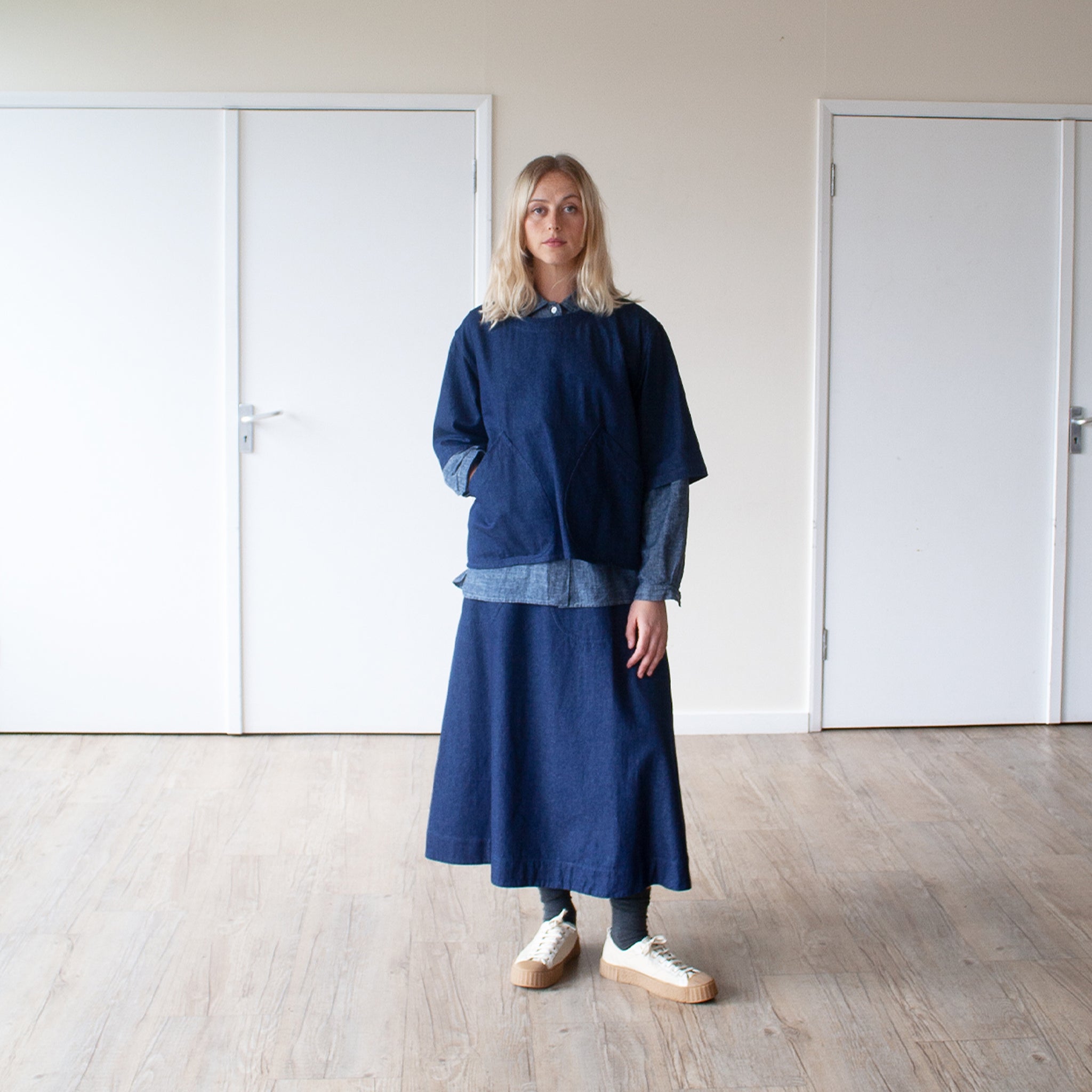 smiling blond woman standing in a sunlit room wearing an indigo denim overtop and skirt facing forward
