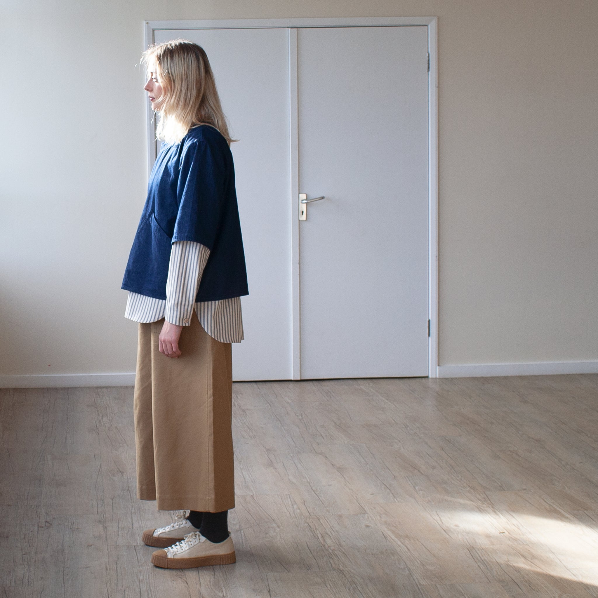 young blond woman standing in a sy=unlit room, side view wearing a tan cotton pair of trousers