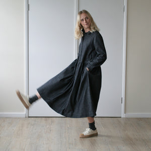 young blonde woman looking forward stands against a cream wall wearing a simple grey denim dress with a gathered waist and long sleeves, she is kicking her leg in the air and has a playful expression on her face