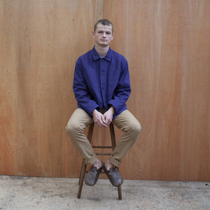 a young man standing against a wooden wall wearing a traditional chore jacket made in a sustainable tan brushed cotton canvas  chore jacket in indigo worn by a young man sitting on a stool
