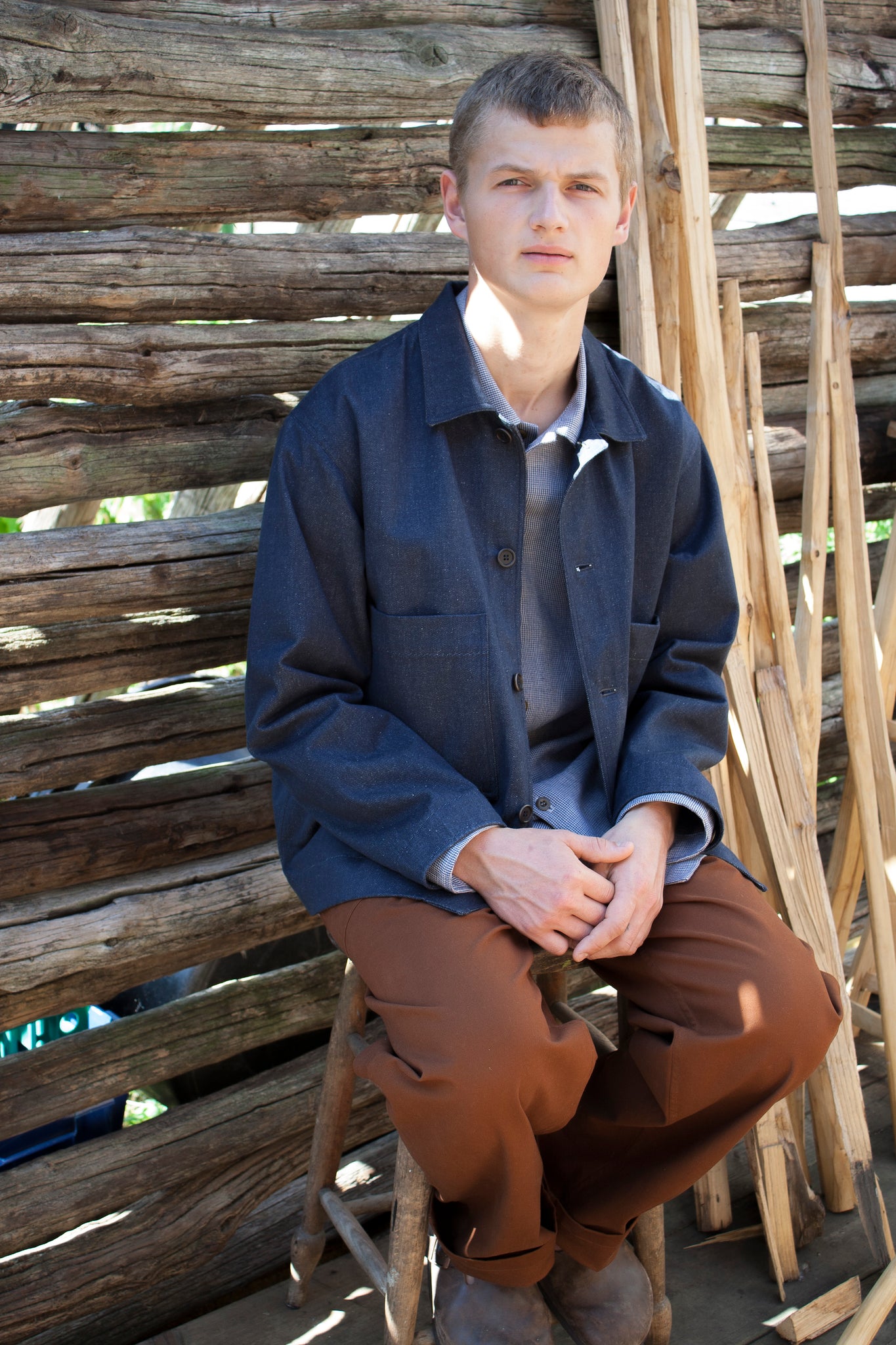 Young man sitting on a stool outside on a sunny day, he is sitting against a wooden wall, and next to some striped wooden poles, he is looking straight at the camera and is wearing a pair of brown trousers and a simple denim workwear jacket