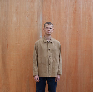 a young man standing against a wooden wall wearing a traditional chore jacket made in a sustainable tan  brushed cotton canvas