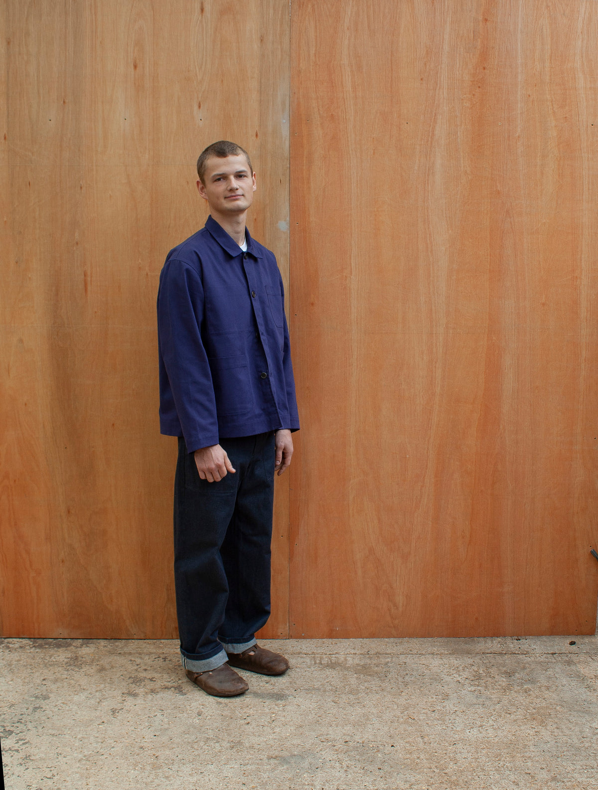 a young man standing against a wooden wall wearing a traditional chore jacket made in an indigo brushed cotton canvas