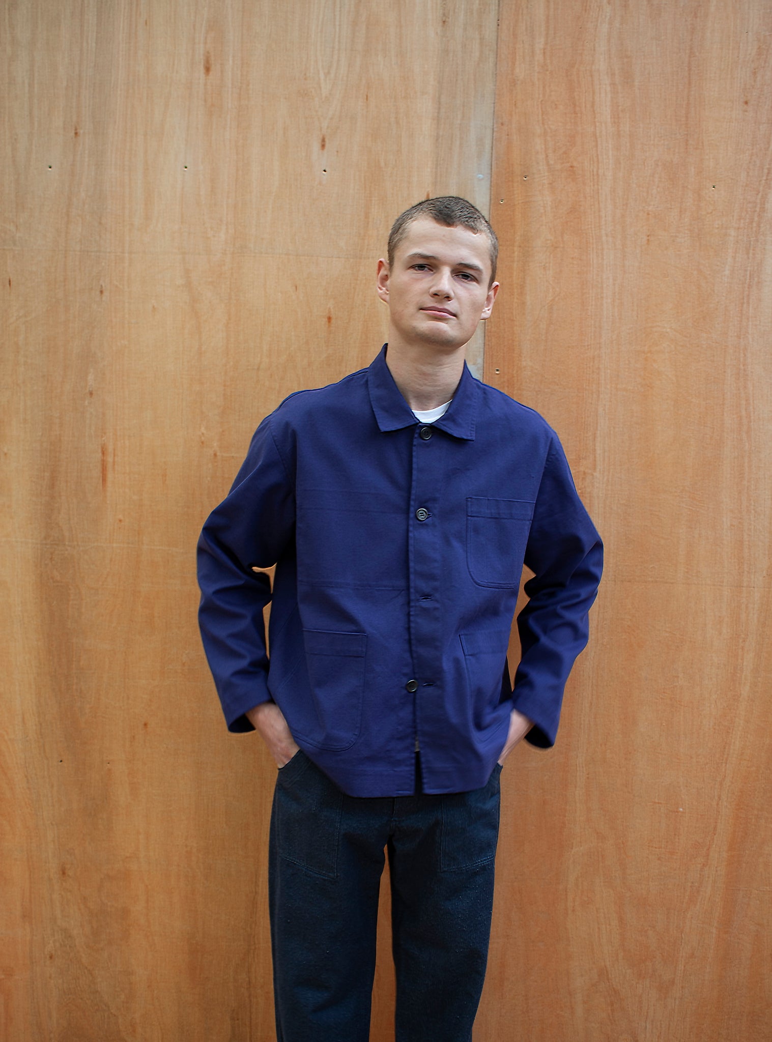 a young man standing against a wooden wall wearing a traditional chore jacket made in an indigo brushed cotton canvas