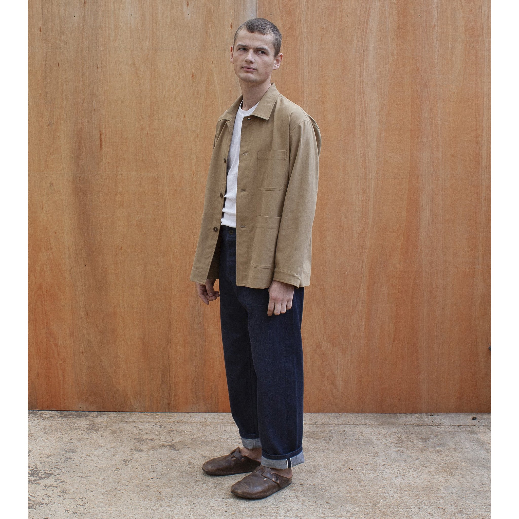 a young man standing against a wooden wall wearing a traditional chore jacket made in a sustainable tan brushed cotton canvas  Edit alt text