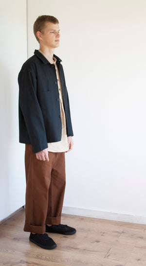 the side view of a young man with short hair, he is standing in the corner of a white room and he is wearing a pair of baggy rust workwear trousers, a grandad shirt and a simple jacket in a soft navy brushed cotton canvas.