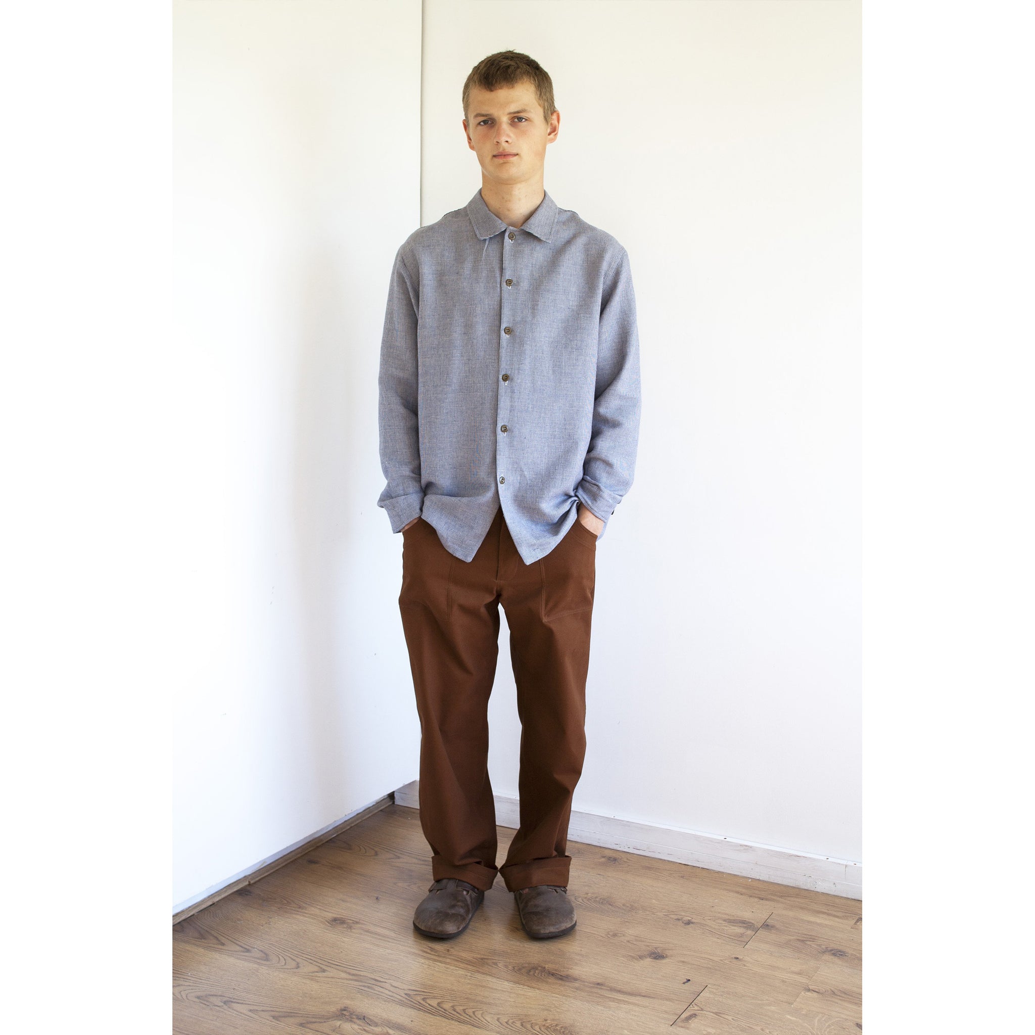young man standing in the corner of a white room, he is looking at the camera and wearing a simple loose fit blue and white linen checked shirt and a pair of rust coloured cotton canvas chore trousers