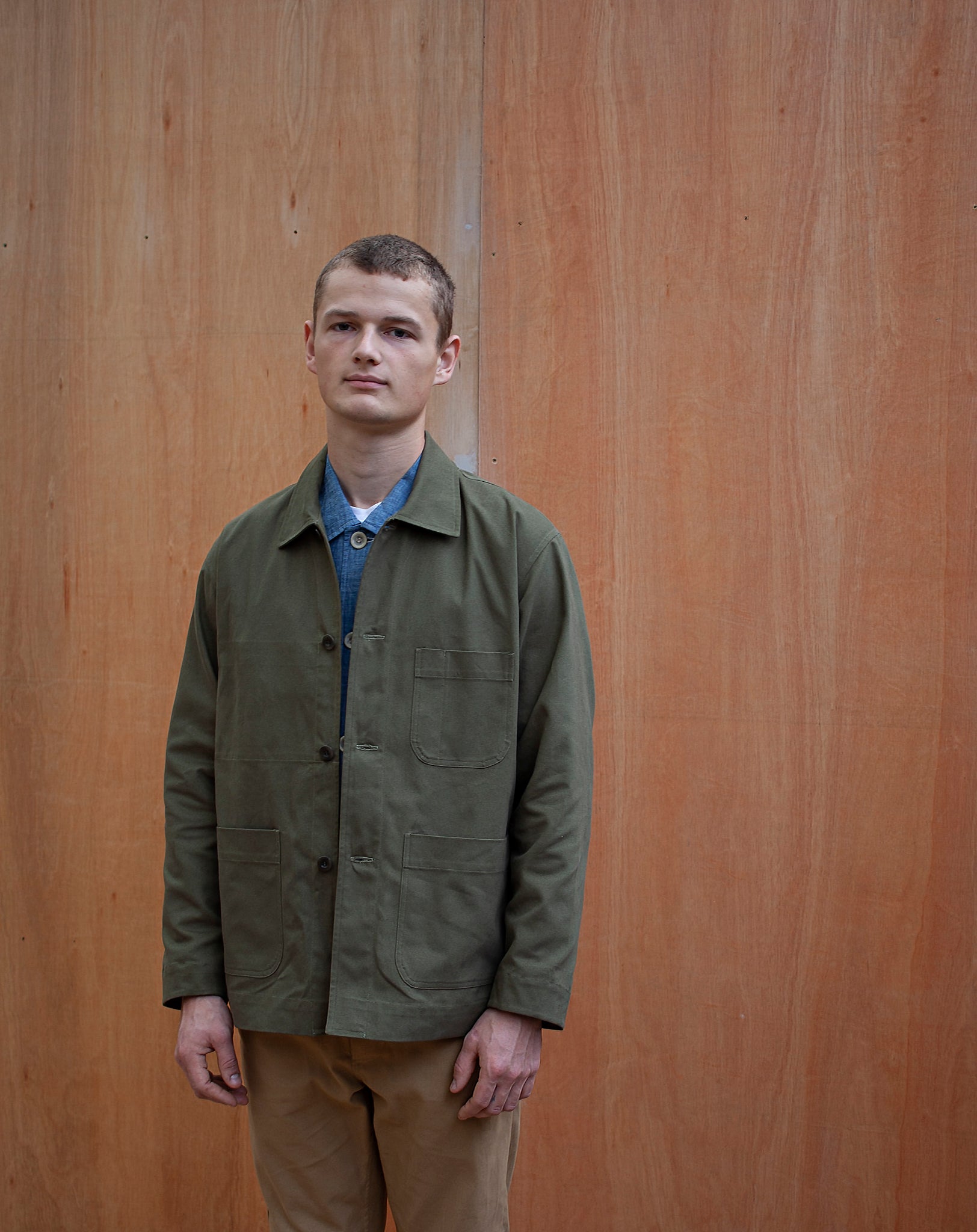 a young man standing against a wooden wall wearing a traditional chore jacket made in a sage green  brushed cotton canvas
