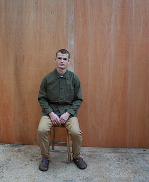 a young man sitting against a wooden wall wearing a traditional chore jacket made in a sage green  brushed cotton canvas