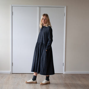 young blonde woman looking forward stands against a cream wall wearing a simple grey denim dress with a gathered waist and long sleeves side view with her hands in her pockets