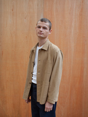 a young man standing against a wooden wall wearing a traditional chore jacket made in a sustainable tan  brushed cotton canvas