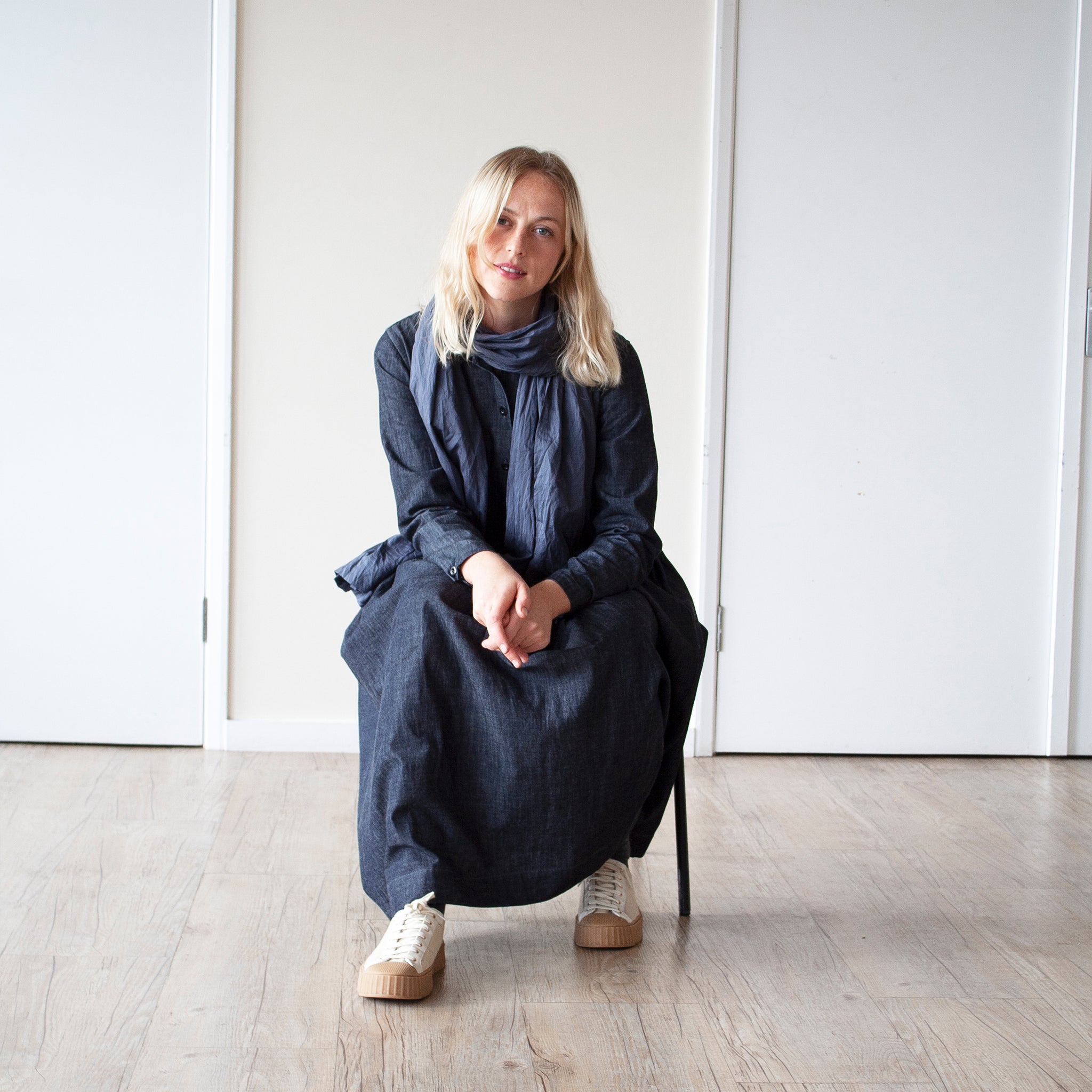 young blonde woman looking forward stands against a cream wall wearing a simple grey denim dress with a gathered waist and long sleeves she is sitting down