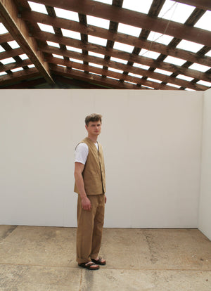 a young man stands sideways to you, wearing a tan coloured waistcoat and matching chore trousers